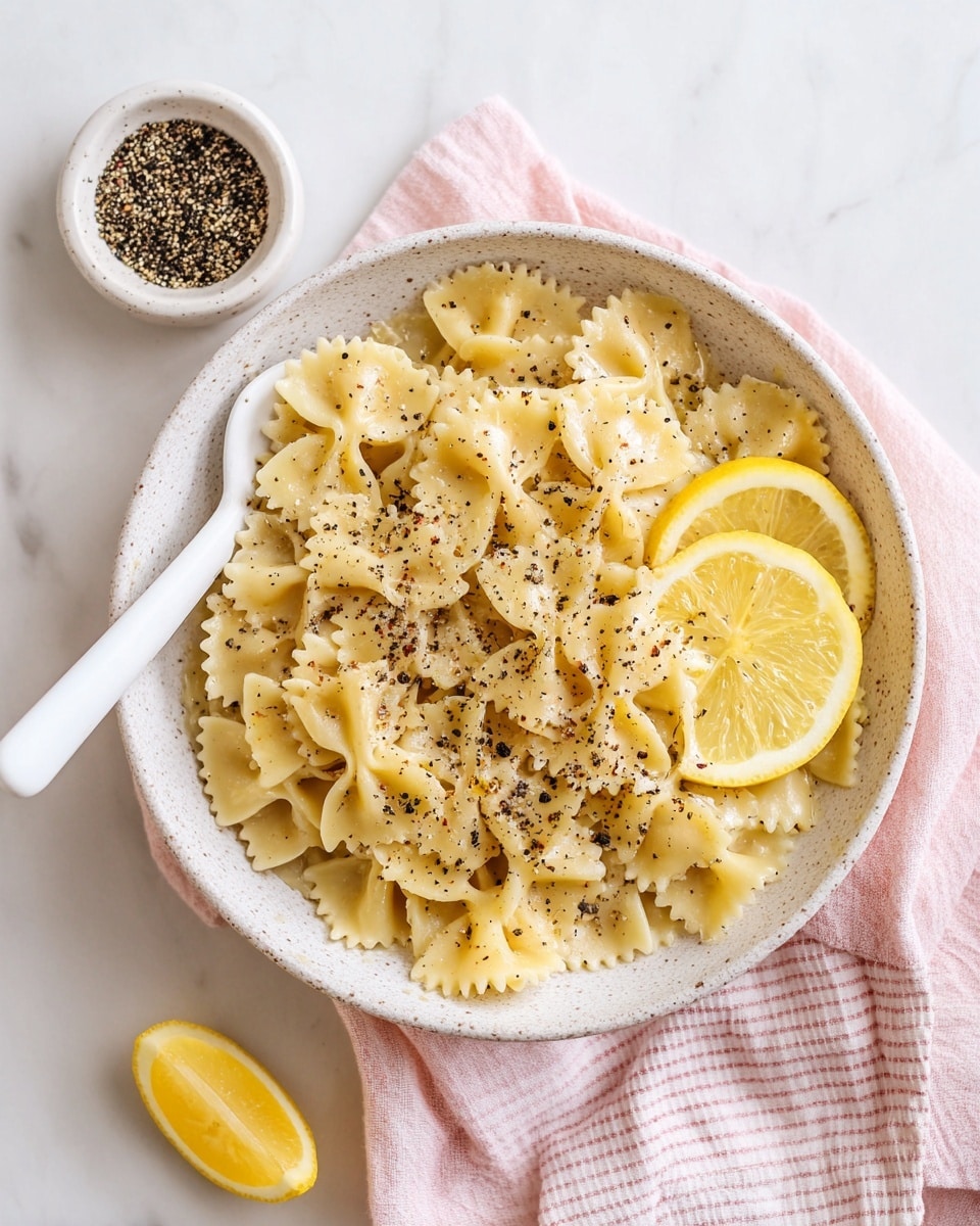 A white speckled bowl filled with a single layer of bowtie pasta coated in a creamy, light yellow sauce, sprinkled with coarse black pepper. Two thin lemon slices rest on the side of the pasta inside the bowl. A white spoon is placed on the left edge of the bowl, which sits atop a soft pink and white striped cloth on a white marbled surface. Nearby, a small white bowl contains more cracked black pepper, and a single lemon wedge is placed on the surface. Photo taken with an iphone --ar 4:5 --v 7