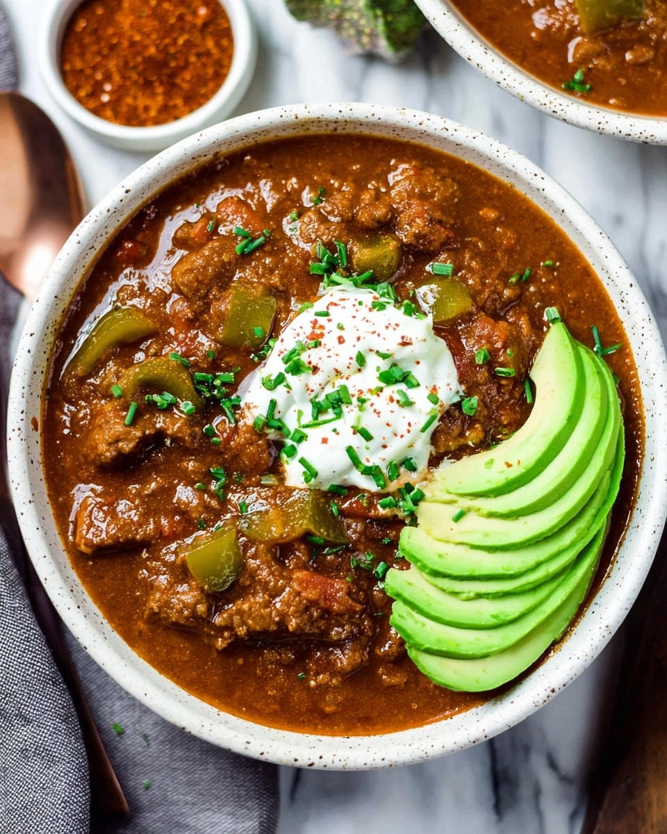 A white speckled bowl filled with thick brown chili, showing chunks of beef and green bell peppers mixed in. On top, there is a dollop of white sour cream sprinkled with green chives and a few pieces of paprika powder. To the right side of the bowl, several slices of bright green avocado fan out, resting on the chili. The bowl is placed on a white marbled surface with a small white dish of reddish-brown seasoning visible nearby. The overall look is warm and hearty with contrasting colors of green, brown, and white. Photo taken with an iphone --ar 4:5 --v 7