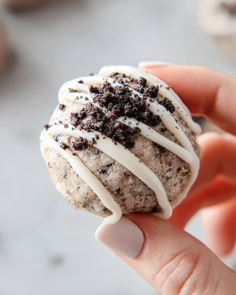 The image shows a small round dessert ball held delicately between a woman's thumb and forefinger against a soft, white marbled background. The dessert has a textured base layer of light brown with visible small dark flecks throughout, giving it a crumbly, cookie-like appearance. On top, there are thin white icing drizzles crossing diagonally, adding a smooth contrast. Finally, dark chocolate cookie crumbs are sprinkled on top, providing a crunchy texture and deeper brown color. The focus is tight on the held dessert, showing fine details of each layer and texture. photo taken with an iphone --ar 4:5 --v 7