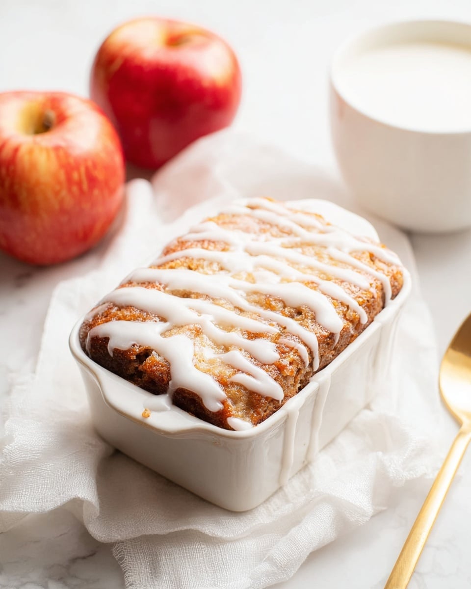 A single square baked dessert sits in a small white ceramic dish on a soft white cloth with a white marbled texture underneath. The dessert has a golden-brown, slightly cracked top layer that looks crispy and textured, with thick white icing drizzled unevenly across the surface. To the left of the dish are three red apples with shiny skins, and on the right side, a shiny gold spoon rests beside a white bowl filled with milk, all placed on the white marbled background. Photo taken with an iphone --ar 4:5 --v 7