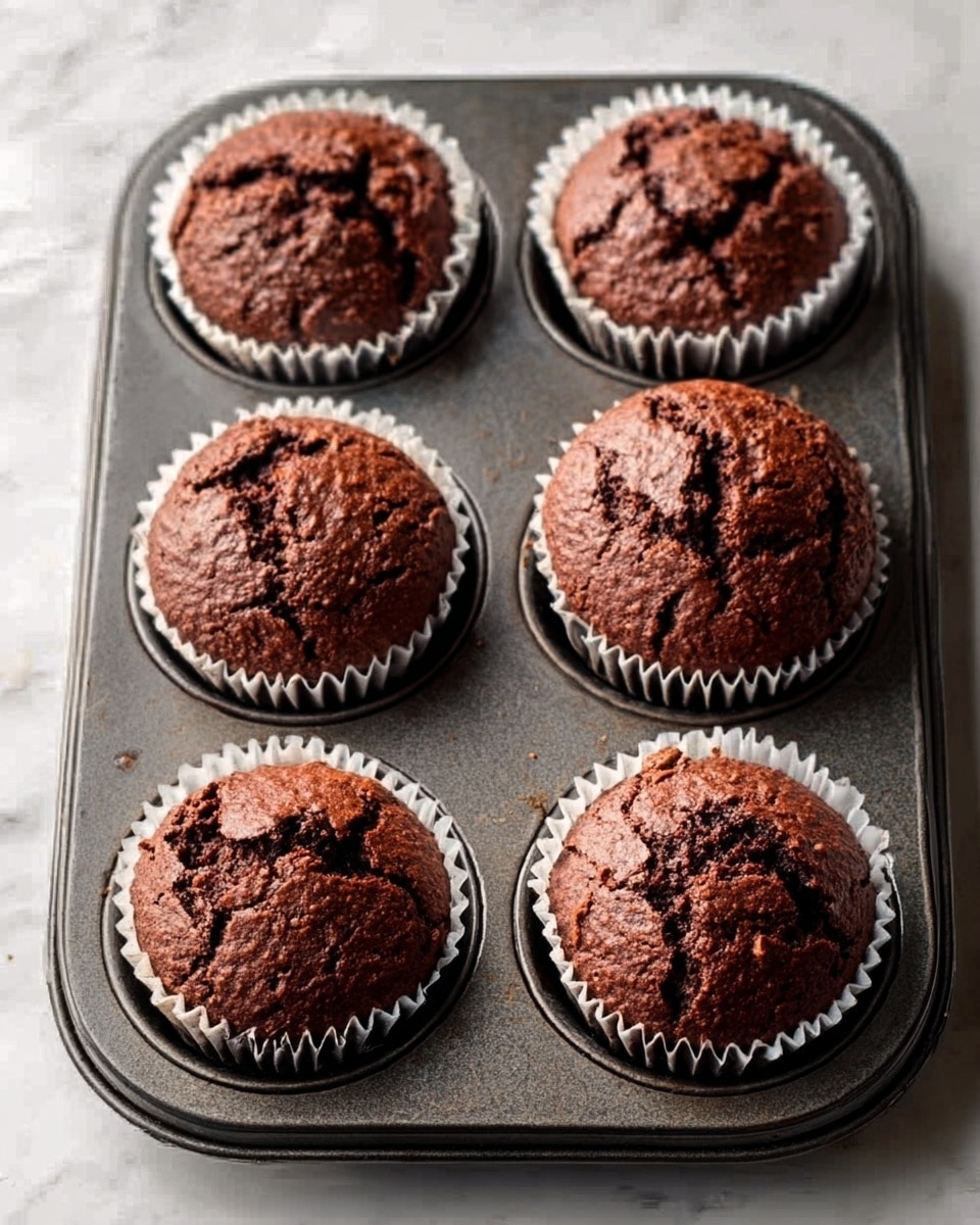 A single chocolate cupcake with a rough, cracked top is placed in the center of a plain white plate. The cupcake liner is light brown with visible ridges, holding a dark brown, moist cake that is topped with small, uneven chocolate sprinkles. The background shows other blurred cupcakes on a white marbled surface, creating a soft and simple setting. The lighting captures the texture of the chocolate cake and sprinkles well. photo taken with an iphone --ar 4:5 --v 7