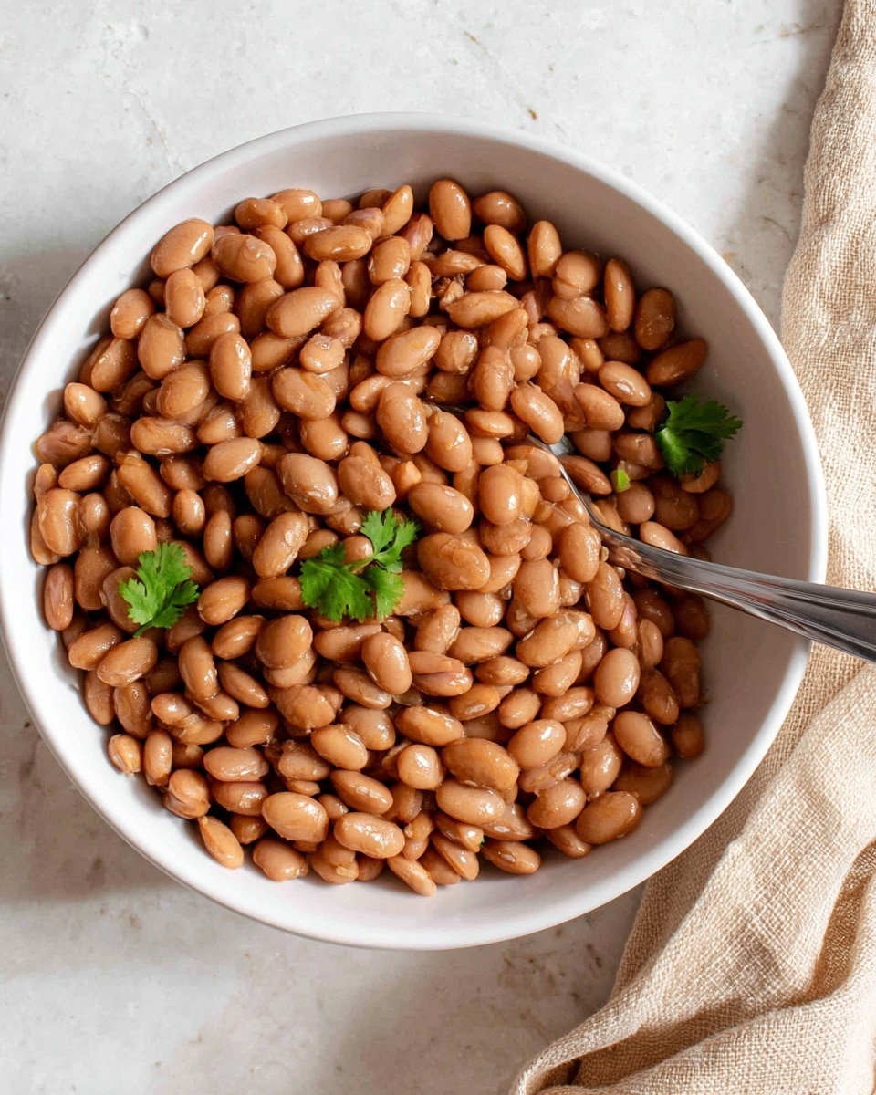 A white bowl filled with a single layer of cooked light brown beans, with a few green cilantro leaves scattered on top and mixed in. A silver spoon is placed inside the bowl on the right side, slightly submerged in the beans. The bowl sits on a white marbled surface, next to a clove of garlic on the bottom left and a piece of green cilantro just below it. A beige cloth is visible in the top right corner, partially in the frame. photo taken with an iphone --ar 4:5 --v 7