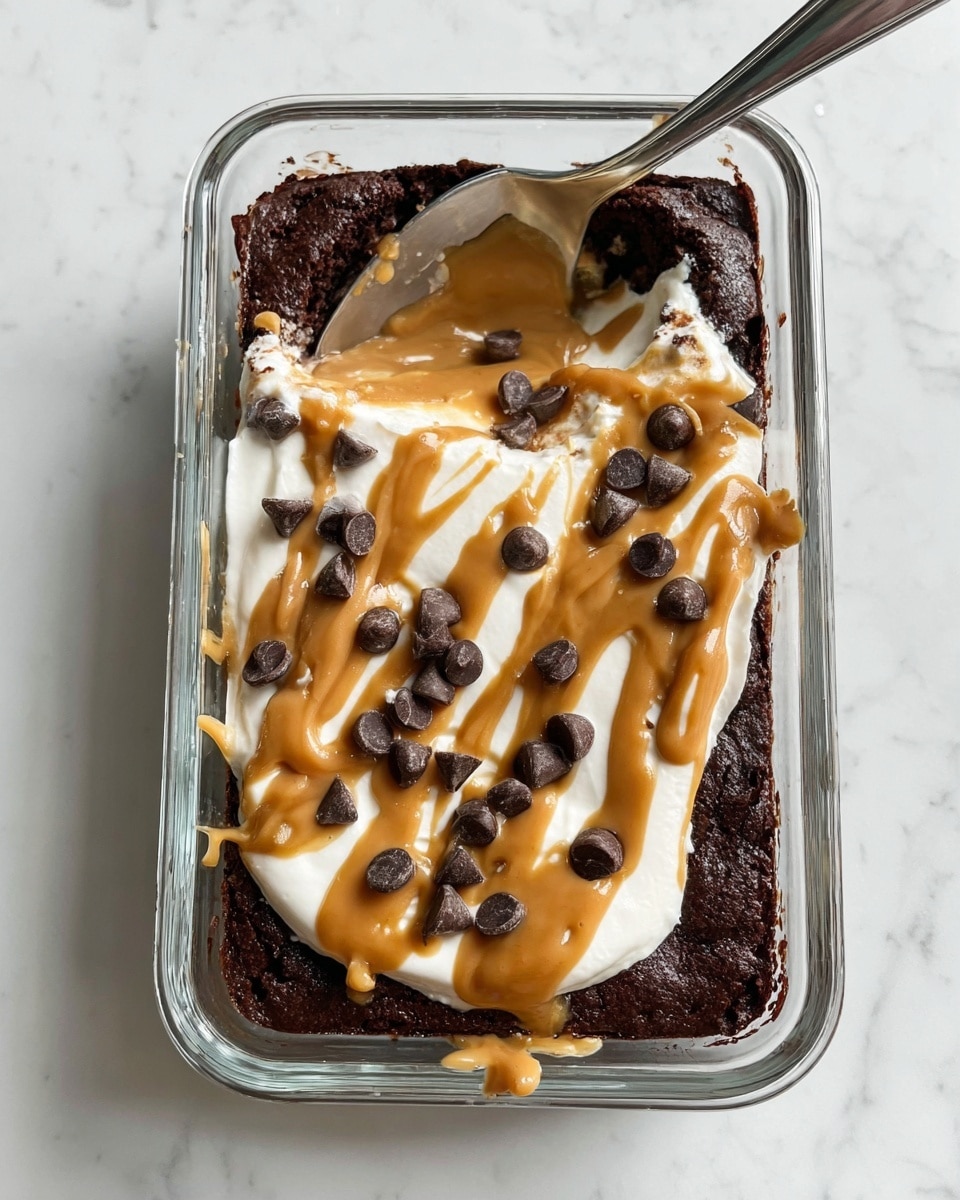 A rectangular, dark brown brownie sits at the bottom of a clear glass dish. On top, there is a smooth white cream layer spread mostly in the center, with thick drizzles of light brown peanut butter sauce flowing over the cream and parts of the brownie in a random pattern. Scattered on top are several glossy, dark chocolate chips. A metal spoon is placed inside the dish on the upper left side. The entire dish is set on a white marbled surface. photo taken with an iphone --ar 4:5 --v 7