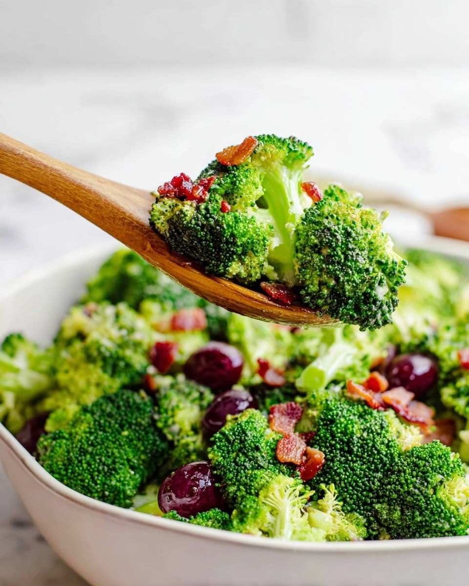 The image shows a fresh broccoli salad with bright green broccoli florets as the main layer, mixed with small pieces of crispy bacon and some dark purple grapes. The broccoli pieces are large and textured, placed in a white bowl on a white marbled surface. A wooden spoon is lifting a portion of the salad, showing a close-up of broccoli with bits of bacon on top, giving a mix of green and reddish-brown colors against the smooth wooden texture of the spoon. Photo taken with an iphone --ar 4:5 --v 7