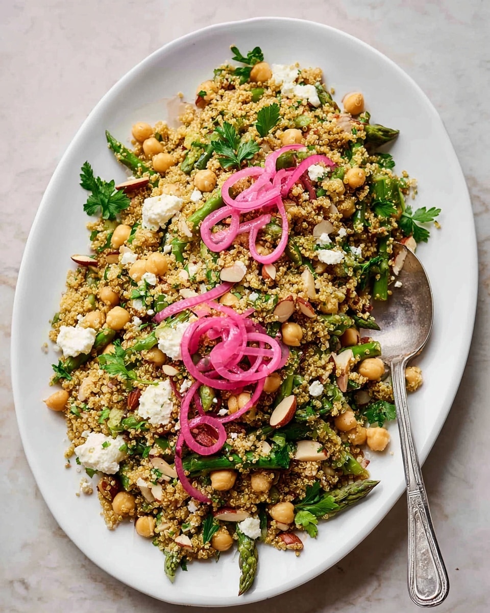 A white oval plate filled with a colorful quinoa salad layered with round, light beige chickpeas and chopped green asparagus pieces. Scattered across the salad are chopped almonds with a light brown appearance and small white crumbles of cheese. Bright pink pickled onion rings are placed in three small clusters on top, adding a pop of color. Fresh green parsley leaves are mixed throughout and placed on one side for garnish. A silver spoon rests on the right side of the plate, containing some of the salad. The plate sits on a white marbled surface. photo taken with an iphone --ar 4:5 --v 7