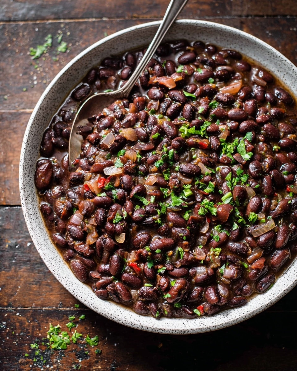 A white speckled bowl filled with cooked black beans mixed with diced onions, small pieces of red pepper, and herbs, all combined in a slightly thick, dark sauce. The beans and vegetables create a textured, chunky layer that almost fills the bowl to the top. Bright green chopped herbs are sprinkled on top, adding fresh color contrast to the deep brown and black tones of the beans. A silver spoon rests inside the bowl, partially submerged in the beans. The bowl is set on a dark wooden surface, with small bits of herbs scattered around. Photo taken with an iphone --ar 4:5 --v 7