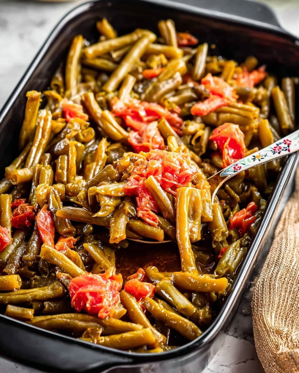 A close-up view of a black rectangular baking dish filled with cooked green beans cut into short pieces, mixed with small chunks of red tomatoes, all in a glossy brown sauce that coats the beans evenly. A silver spoon with a floral pattern on the handle is pulling some of the beans and tomato pieces from the dish. The dish sits on a white marbled surface, with a tan-colored textured cloth partially visible on the right side. The photo taken with an iphone --ar 4:5 --v 7