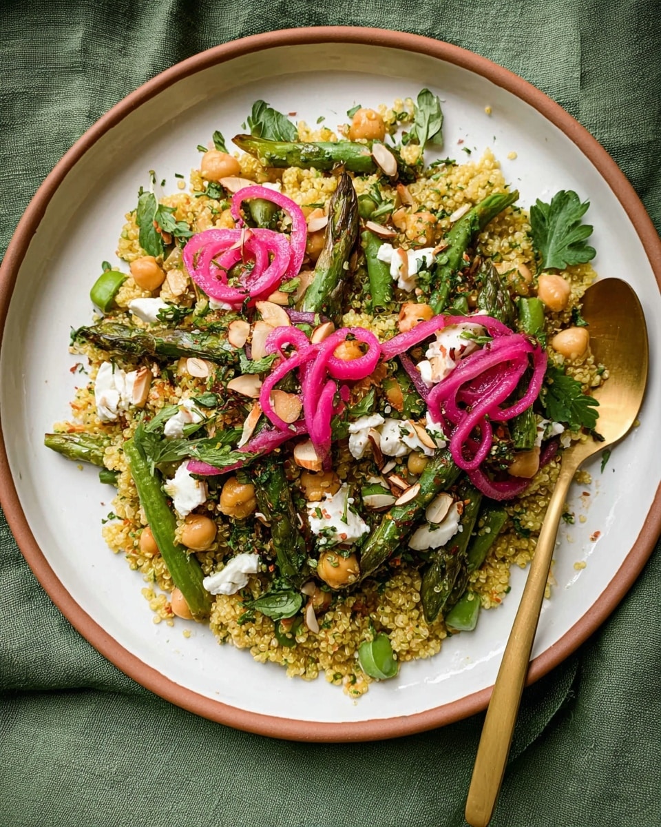 A white plate with a thin brown rim holds a colorful salad with several layers: a base of yellow quinoa mixed with green herbs and small chickpeas, topped with green snap peas and asparagus pieces, scattered beige almond chunks, and white crumbles that look like cheese. Bright pink pickled onion curls sit on top in two places, along with some fresh green parsley leaves scattered around. A gold spoon rests on the edge of the plate on the right side. The plate is set on a green cloth with a subtle texture. Photo taken with an iphone --ar 4:5 --v 7