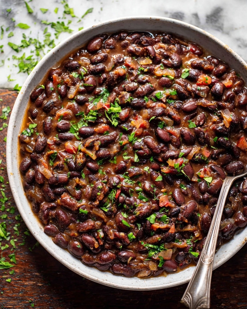 A close-up of a white bowl full of cooked black beans in a thick brown sauce mixed with small pieces of onions, tomatoes, and garnished with chopped green herbs, with a silver spoon resting on the right side of the bowl. The bowl is placed on a white marbled texture. Some scattered green herbs are on the surface around the bowl. photo taken with an iphone --ar 4:5 --v 7