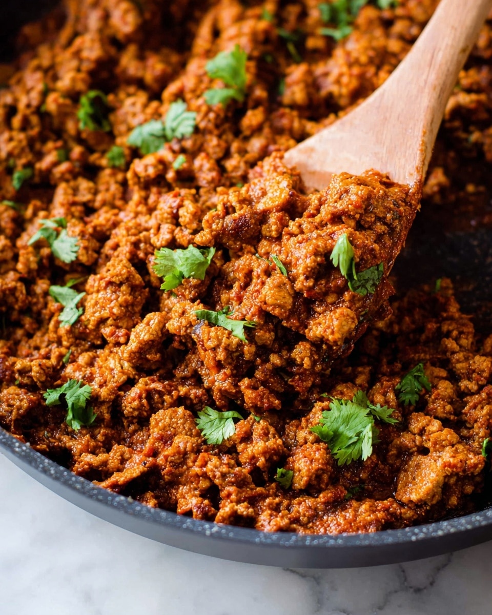 A close-up view of a pan filled with cooked ground meat mixed with rich, reddish-brown sauce, showing textured chunks coated in spices. Small bright green coriander leaves are scattered on top, adding pops of color. A wooden spoon scoops a portion, lifting the mixture with a slightly crispy, browned surface on some pieces. The pan edges show a smooth, dark surface, all set against a white marbled texture. Photo taken with an iphone --ar 4:5 --v 7