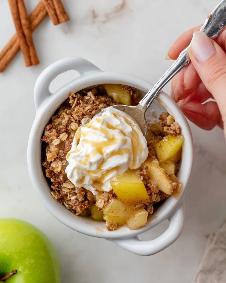 A white small round bowl with two handles holds a warm apple crisp dessert with visible chunks of cooked yellow apple mixed with a crumbly, golden-brown oat and cinnamon topping. On top, there is a swirl of creamy white whipped cream drizzled with light golden syrup. A silver spoon scoops into the dessert, held by a woman's hand. The bowl is placed on a white marbled surface with two cinnamon sticks in the top left corner and a green apple near the bottom left edge of the image. photo taken with an iphone --ar 4:5 --v 7