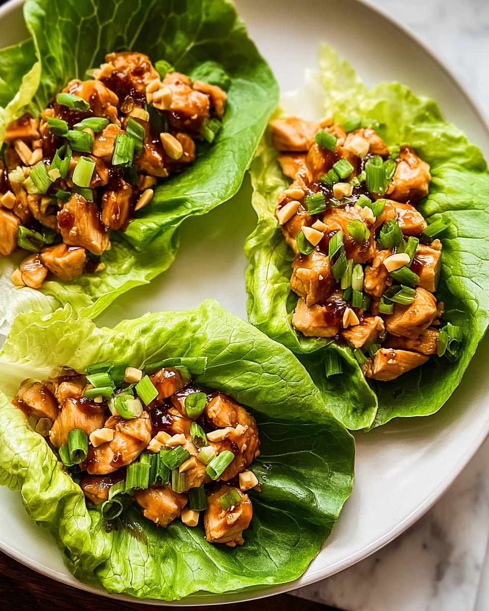 This image shows three lettuce wraps placed on a white plate with a dark texture, sitting on a white marbled surface. Each wrap has a fresh, bright green large lettuce leaf as the base layer, cradling a layer of small, cooked chicken pieces coated in a shiny, rich brown sauce. On top of the chicken, chopped green onions add a pop of lighter green color, mixed with small bits of peanuts, adding crunch and texture. The wraps have a fresh, vibrant look with a crisp and glossy finish on the lettuce and chicken layers. Photo taken with an iphone --ar 4:5 --v 7