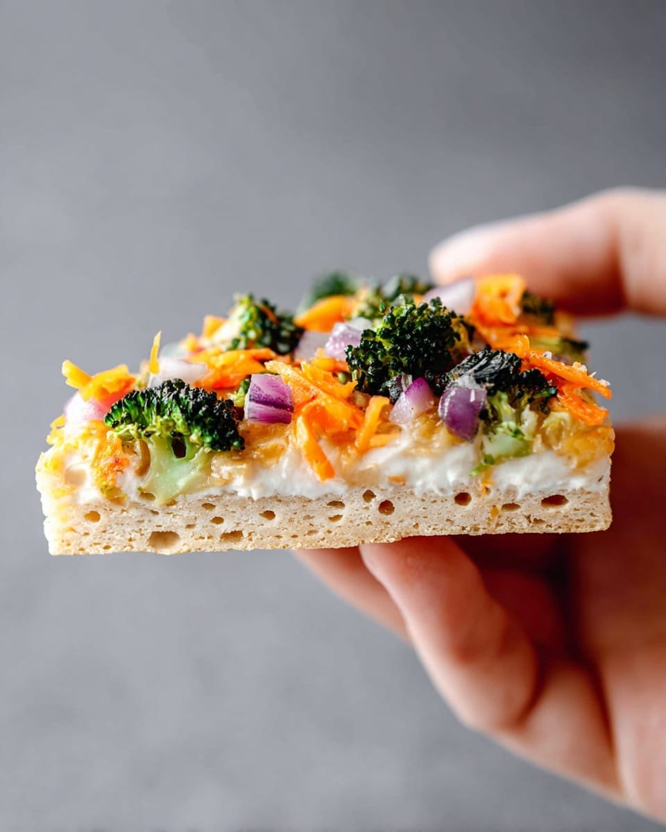 A close-up slice of a vegan cracker pizza is held by a woman's hand against a plain gray background. The pizza has three visible layers: the bottom layer is a light beige, airy cracker base with small holes; the middle layer is a smooth, creamy white spread; the top layer is a mix of small bright orange shredded carrots, dark green broccoli florets, and tiny pieces of purple-red onion, all scattered unevenly across the surface. The textures vary from the crunchy cracker base to the soft spread and fresh vegetables. Photo taken with an iphone --ar 4:5 --v 7