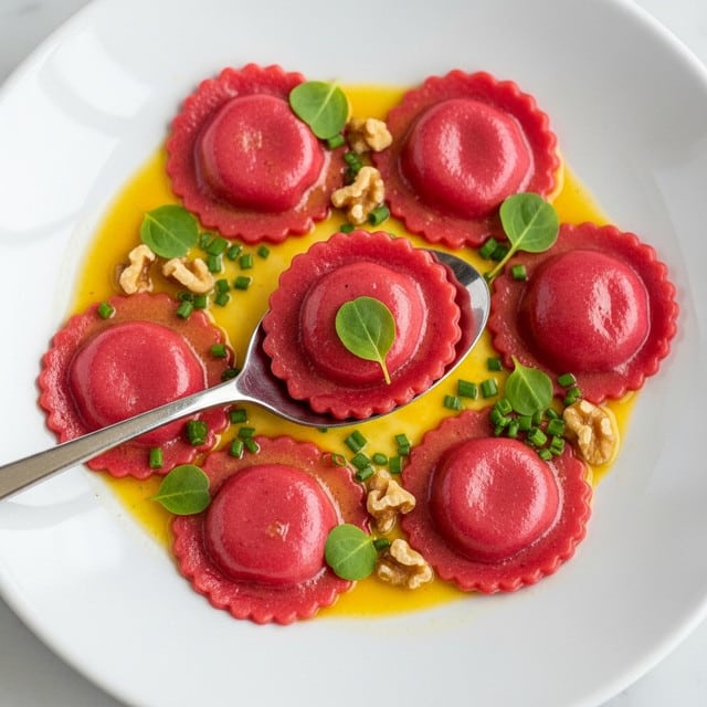 A white plate holds about twelve pink ravioli arranged in a loose circle, each ravioli round with crimped edges and a smooth, slightly shiny pink surface. The ravioli float in a light orange-yellow butter sauce with a glossy texture. Scattered on top and around the ravioli are small green herbs like dill and mint, candied walnut pieces adding a warm brown touch, and some green chive-like sticks. A silver spoon lifts one ravioli in the center, showing the pink color and texture up close, with tiny herb leaves on top. The whole scene is on a white marbled surface. photo taken with an iphone --ar 4:5 --v 7