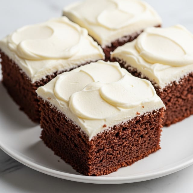 Three square chocolate cakes with a thick layer of smooth, creamy white frosting on top are placed close together on a simple white plate. The cakes have a rich, dark brown color with a soft and moist texture that contrasts with the light, fluffy frosting. The frosting is evenly spread with gentle swirls visible on each piece. The plate is set on a white marbled surface with soft natural light highlighting the smoothness of the frosting and the moist texture of the cake. photo taken with an iphone --ar 4:5 --v 7