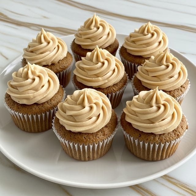 A white plate holds eight cupcakes arranged in a close grouping on a white marbled surface with gold streaks. Each cupcake has a light brown base with a slightly rough texture and white paper liners. On top of each is a single large swirl of smooth, creamy frosting in a light tan color, shaped like a soft peak with gentle curves and ridges. The frosting looks rich and thick, sitting neatly centered on each cupcake. photo taken with an iphone --ar 4:5 --v 7