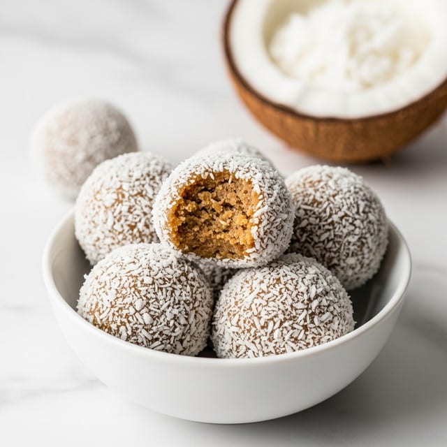 A white bowl holds five round dessert balls, each coated with white coconut flakes on the outside, giving a textured white layer over a light brown inner layer that is visible on one ball with a bite taken out, showing a crumbly and moist texture. In the background, there is a blurred half coconut shell with white flesh, all set on a clean white marbled surface. The focus is on the bowl and the front dessert balls, creating a simple and fresh look. photo taken with an iphone --ar 4:5 --v 7