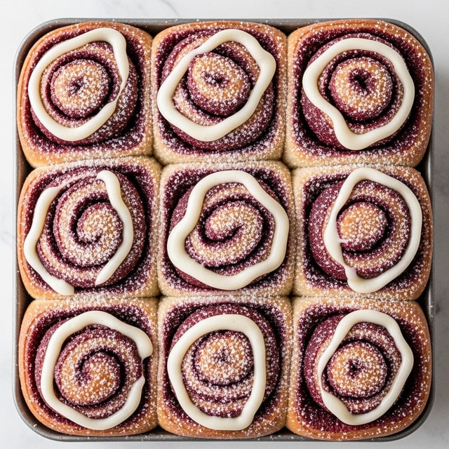 The image shows a close-up of nine red velvet cinnamon rolls tightly packed in a metal baking pan. Each roll has a golden-brown dough base visible at the edges, layered with a deep red, smooth, and slightly textured filling spiraled from the center to the outer edge. On top, there is a thick, creamy white frosting swirled over the rolls in a circular pattern that doesn't completely cover the red filling, allowing the colors to contrast. The rolls are dusted with small white powdered sugar particles that sparkle on the frosting and dough. The pan is placed on a white marbled surface with a soft light highlighting the texture and colors vividly, photo taken with an iphone --ar 4:5 --v 7