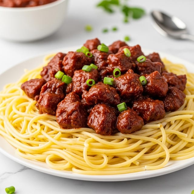 A white plate holds a bed of light yellow spaghetti noodles, topped with chunky pieces of meat covered in a thick, shiny reddish-brown sauce. The meat is garnished with small, bright green chopped herbs scattered on top. In the background, there is another similar plate slightly out of focus, and a white bowl with red sauce and a spoon inside. The scene is set on a white marbled surface with some green herbs loosely placed around. photo taken with an iphone --ar 4:5 --v 7