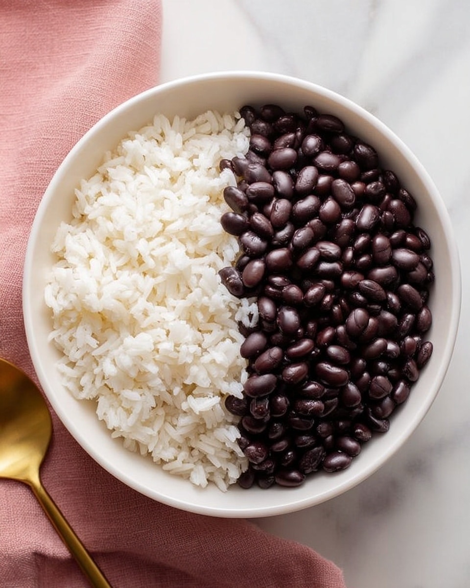 A white bowl filled with two equal halves, one side packed with fluffy white rice showing its soft grains and the other side filled with shiny, dark black beans that contrast against the rice. The bowl sits on a pink cloth on top of a white marbled surface, and a golden spoon is placed nearby, adding warmth to the scene. The photo taken with an iphone --ar 4:5 --v 7