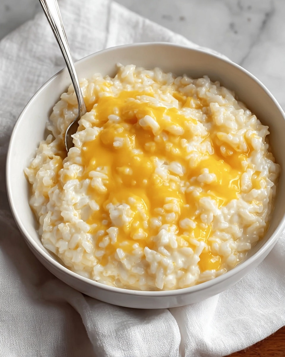 A close-up view of a white bowl filled with creamy cooked rice topped with melted bright yellow cheese. The rice layer is soft and textured with individual grains visible, while the cheese melts smoothly over the top in an uneven spread. A metal spoon rests inside the bowl on the right side, slightly digging into the rice and cheese. The bowl sits on a white cloth on a wooden surface, though the background is changed to a white marbled texture. photo taken with an iphone --ar 4:5 --v 7