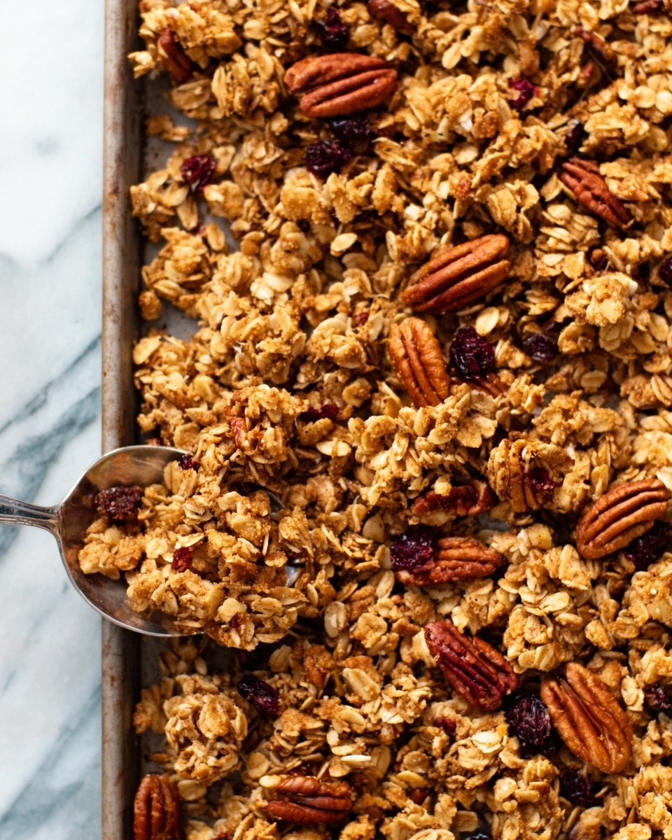 The image shows a close-up of a baking tray filled with a golden brown granola mix. The granola is in rough clusters with visible oats and large, glossy pecans scattered evenly on top. The granola clusters have a crunchy texture with some darker brown spots from baking. The tray is lined with white parchment paper, all set on a white marbled surface. Photo taken with an iphone --ar 4:5 --v 7