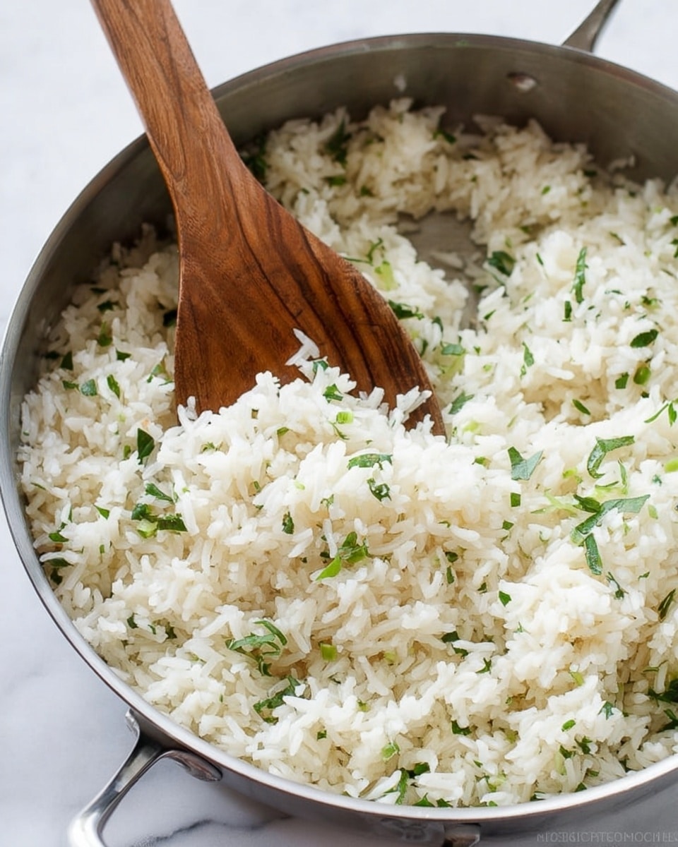 The image shows a close-up of cooked white rice mixed with small green herb pieces, possibly cilantro or parsley, in a shiny silver pan. The rice is fluffy with separate grains visible throughout, and the green herbs are scattered evenly inside. A light wooden spoon is placed upright in the pan, partially covered by the rice, adding a natural texture contrast against the metal. The pan handle is also visible at the bottom, emphasizing the focus on the rice mixture inside. The background features a soft white marbled texture. photo taken with an iphone --ar 4:5 --v 7