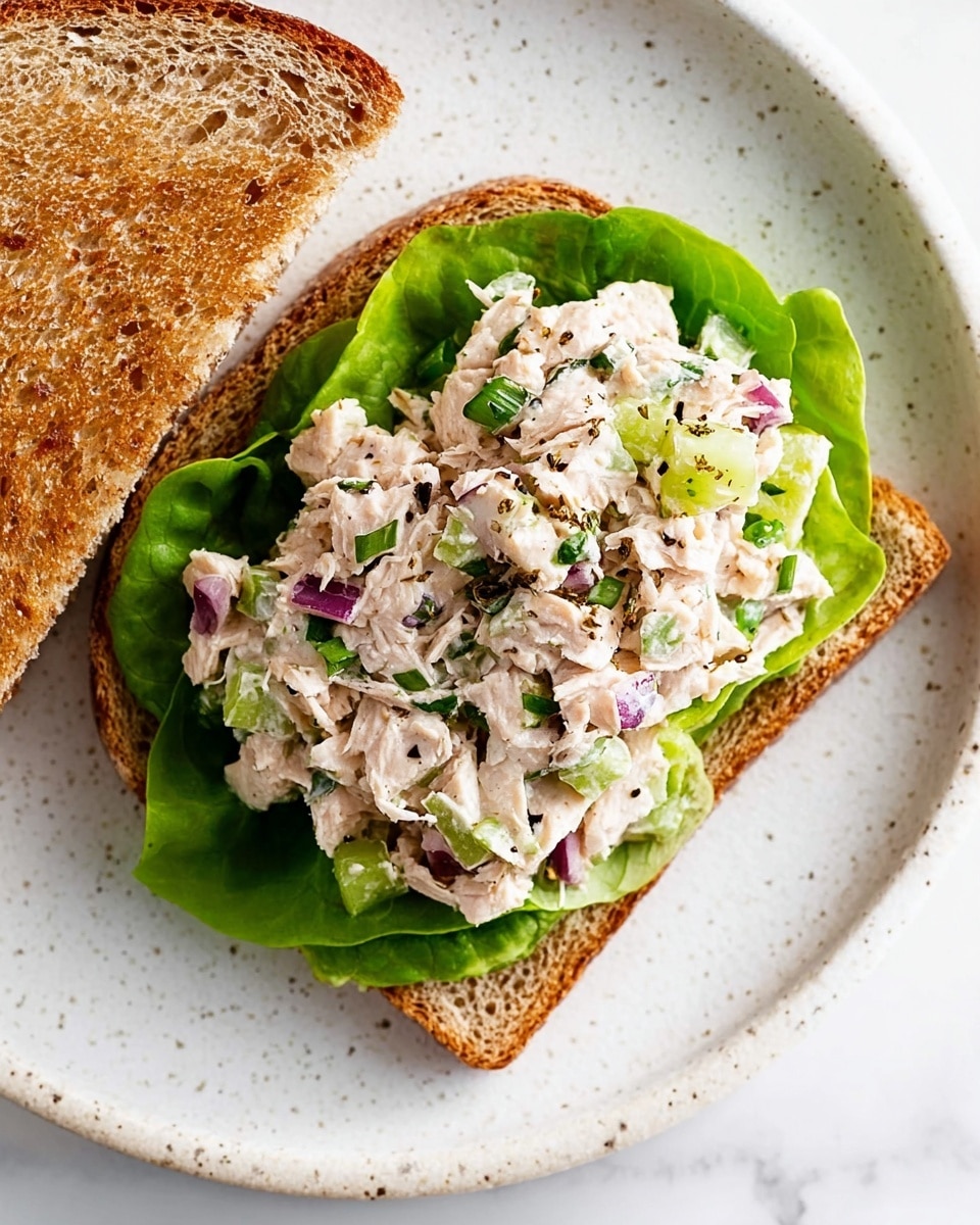 A close-up of an open sandwich on a slice of brown toasted bread placed on a white plate with a speckled edge, set on a white marbled surface. The sandwich has three layers: the bottom layer is a green leafy lettuce with a fresh, crisp texture; the second layer is a chunky mix of light pinkish-white tuna salad with visible pieces of diced red onion, green celery, and chopped herbs, sprinkled lightly with black pepper; the top layer is another slice of toasted brown bread placed slightly to the side, showing its soft texture inside. The colors show a natural, fresh look with contrast between the green, purple, and light tuna tones. Photo taken with an iphone --ar 4:5 --v 7