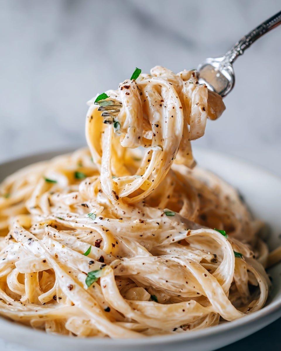 A close-up view of creamy fettuccine pasta coated in a smooth, pale beige sauce with slight specks of black pepper and small bits of green herbs scattered on top. The tangled noodles are lifted by a pair of silver forks, showing the pasta’s soft and silky texture. The whole scene has a creamy, rich look with the pasta strands overlapping closely. The soft lighting highlights the sauce's silkiness and the faint green herb pieces that add a touch of color. The dish is set against a white marbled textured background. Photo taken with an iphone --ar 4:5 --v 7