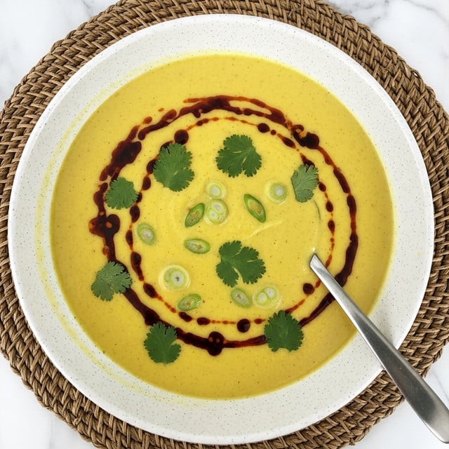A creamy yellow soup fills a white speckled bowl placed on a woven mat; the smooth soup surface is dotted with dark red oil swirls and small green herb pieces scattered evenly across the top. Thin slices of green onion float gently, adding fresh green highlights. A silver spoon rests inside the bowl, partially submerged in the soup, angled toward the front right. The background is a white marbled texture. photo taken with an iphone --ar 4:5 --v 7