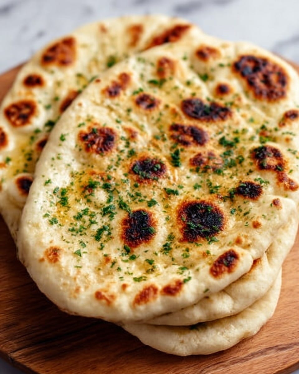Two pieces of naan bread stacked on top of each other on a wooden board, each naan showing a soft, slightly bubbly texture with golden brown and dark charred spots. The top naan is sprinkled with finely chopped green herbs, adding a fresh look. The edges are slightly thicker and puffier, with a light cream color blending into the toasted brown areas. In the background, there is a white marbled surface. photo taken with an iphone --ar 4:5 --v 7