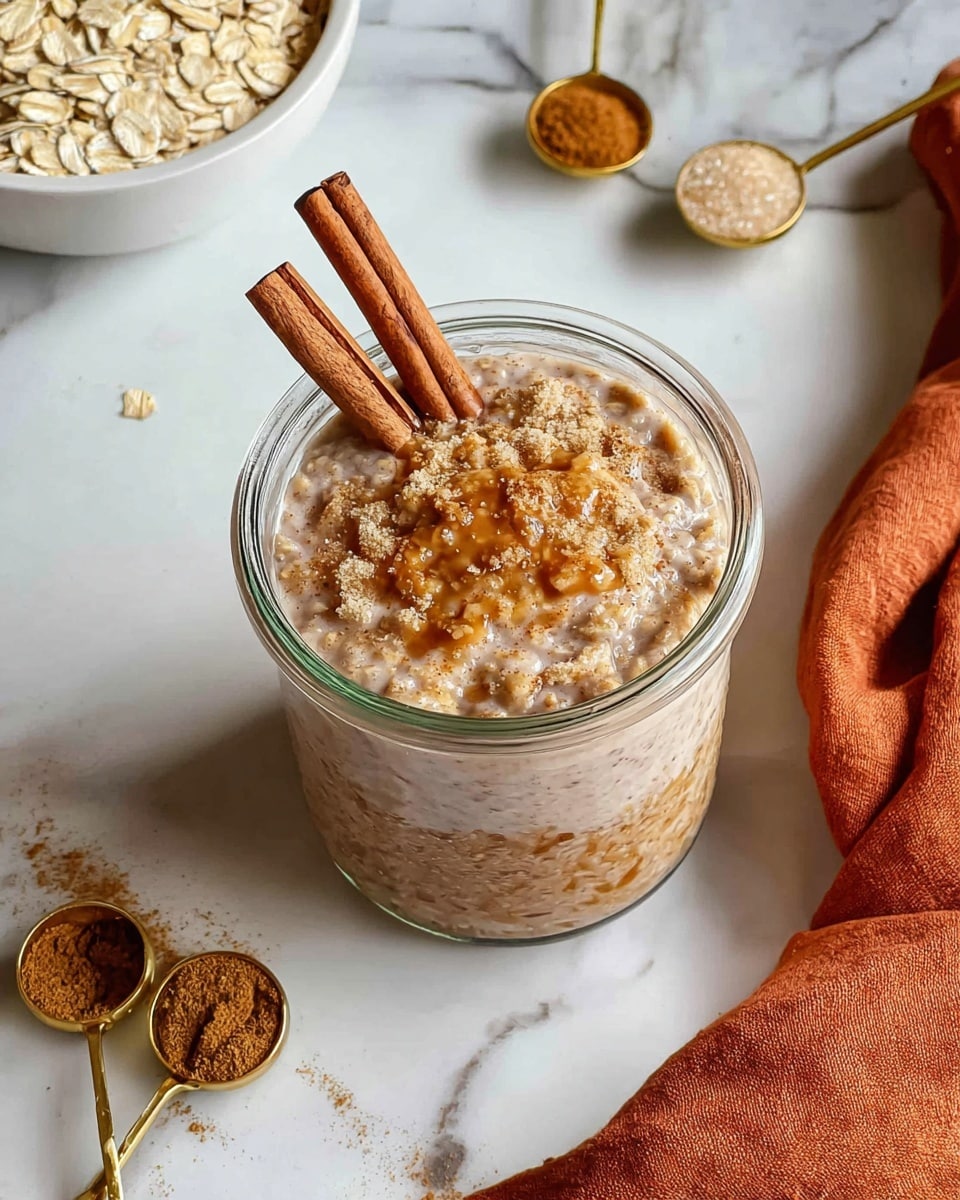 A clear glass jar filled with three layers of oatmeal mixture topped with visible whole oats and a slightly frothy surface, with two cinnamon sticks placed diagonally on top. Around the jar on a white marbled surface are two gold-colored spoons, one filled with ground cinnamon powder and the other with brown sugar, and a white bowl filled with raw oats positioned at the top left. An orange cloth is casually placed on the top right corner. Photo taken with an iphone --ar 4:5 --v 7