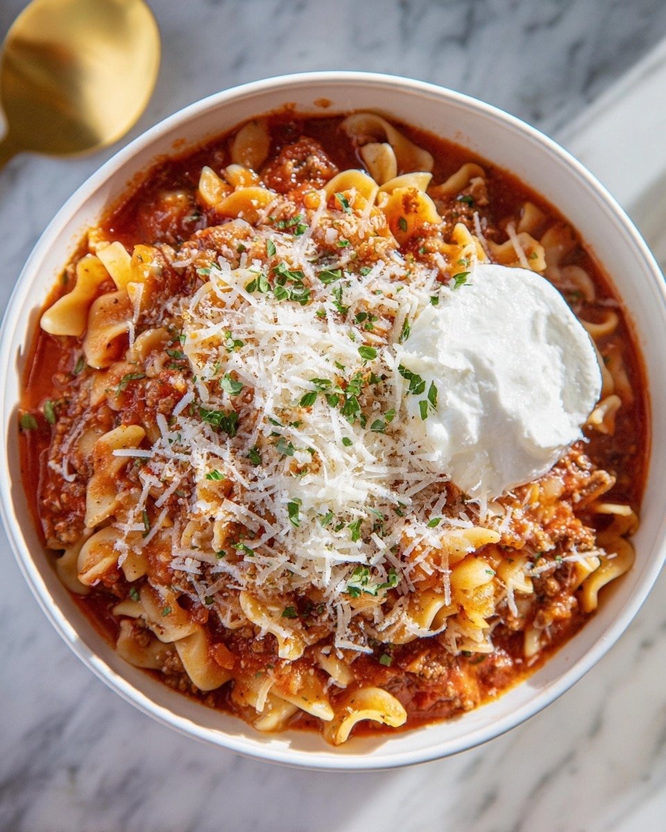 A white bowl filled with layered pasta dish, starting with a soft, curvy pasta base in pale yellow mixed with rich, red tomato meat sauce and scattered small meat pieces. On top, a layer of finely shredded white cheese is spread evenly, mixed with small green herb bits. The dish is finished with a large scoop of white creamy dollop placed on one side. The bowl sits on a white marbled surface with a glimpse of a golden spoon beside it. photo taken with an iphone --ar 4:5 --v 7
