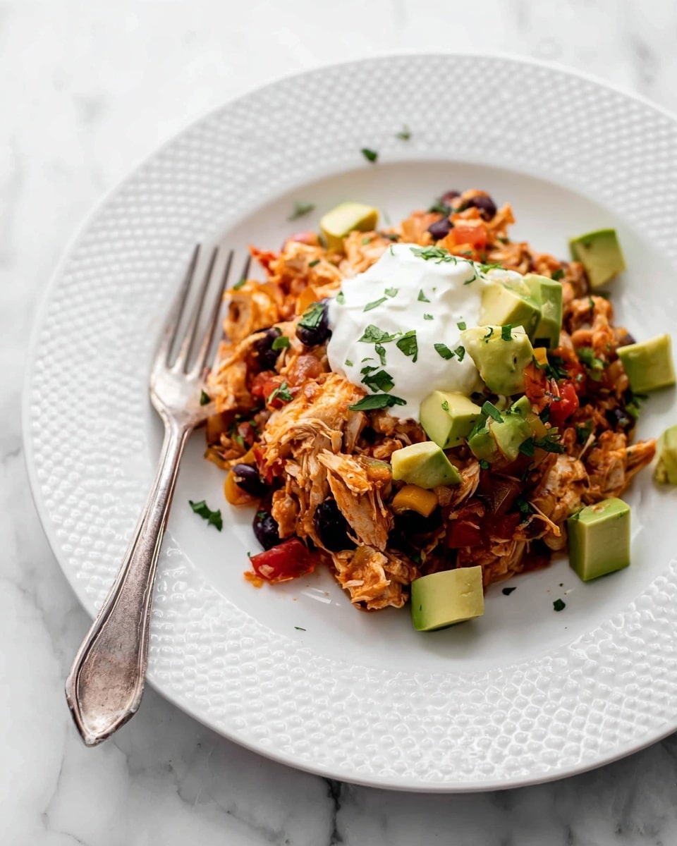 The dish is served on a white plate with ridged edges, sitting on a white marbled surface. The main layer consists of shredded chicken mixed with black beans, corn, and small diced red and yellow bell peppers, all combined with rice. Scattered on top and around this base are chunks of light green avocado. The whole mixture is garnished with chopped green herbs, and in the center, there is a generous dollop of white sour cream. A silver fork rests on the left side of the plate near the food. Photo taken with an iphone --ar 4:5 --v 7