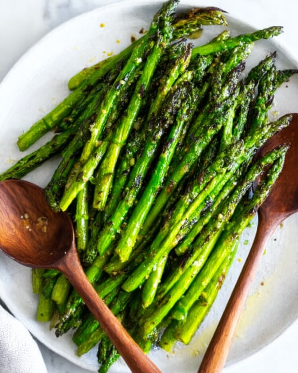 The image shows a white round plate filled with cooked green asparagus spears neatly arranged side by side, all pointing in the same direction. The asparagus has a slightly shiny and tender look with some light grill marks or slight browning, showing they are cooked but still fresh and bright green. Two wooden utensils, a fork and a spoon, are placed on the plate, one on each side of the asparagus. The plate is set on a white marbled surface, adding a clean and elegant background. photo taken with an iphone --ar 4:5 --v 7