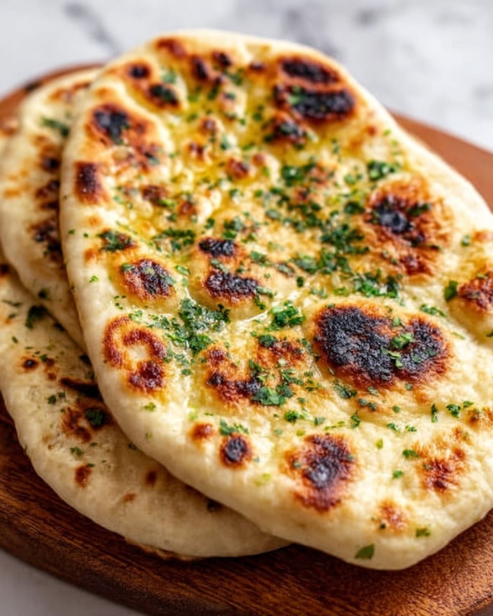 Two round naan breads stacked slightly off-center on a white marbled surface, each with a golden-brown, bubbly texture and dark charred spots on top. The surface of the naan is sprinkled with finely chopped green herbs, adding a fresh look to the warm, soft bread. The naan’s edges are rounded and puffed, showing a light, airy inside. photo taken with an iphone --ar 4:5 --v 7