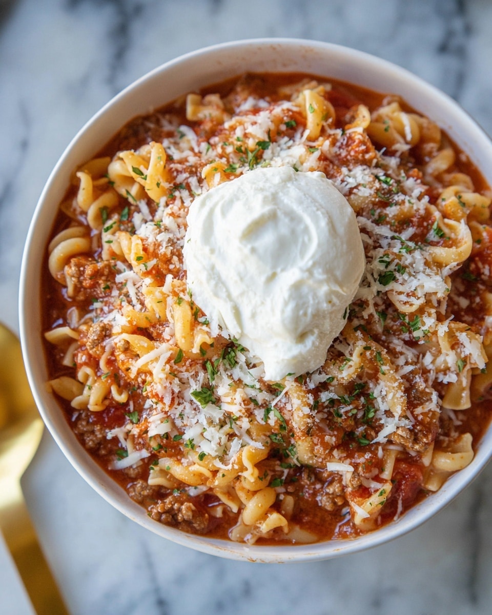 A white bowl filled with a pasta dish showing two clear layers: the bottom layer is soft, curly pasta mixed with a thick red tomato sauce and small bits of cooked ground meat, while the top layer is sprinkled with grated white cheese and small pieces of green herbs for color contrast. On the top right side, there is a large dollop of smooth, white cream. The bowl sits on a white marbled surface with a blurred golden spoon handle visible to the side. photo taken with an iphone --ar 4:5 --v 7