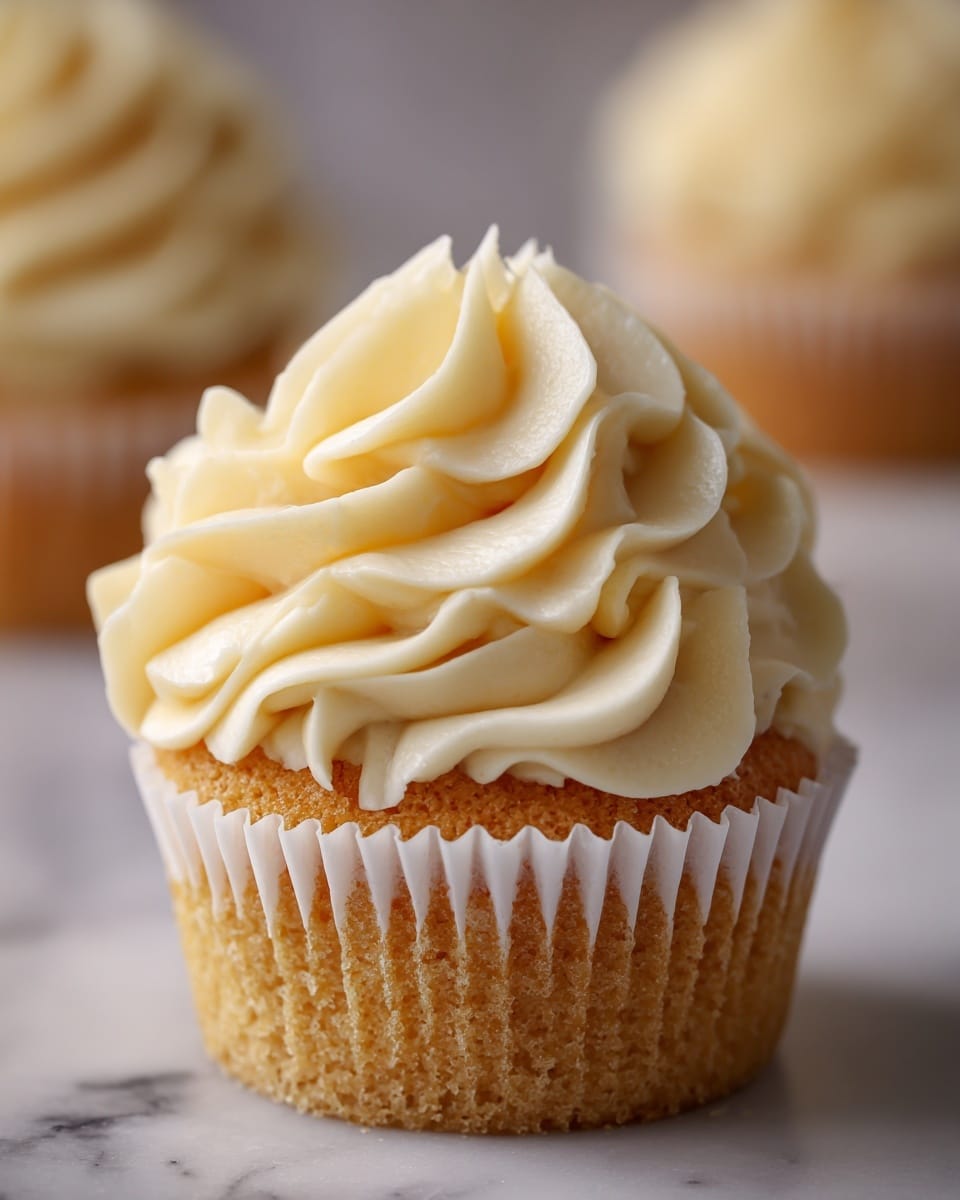 A close-up view of a single cupcake in a white paper liner sitting on a white marbled surface. The cupcake has one visible layer of light golden cake at the bottom, topped with a thick, creamy swirl of smooth pale yellow frosting that is piped in large, soft peaks, creating a textured and fluffy look. The background is softly blurred with a hint of another cupcake, keeping the focus on the detailed frosting and cake. Photo taken with an iphone --ar 4:5 --v 7