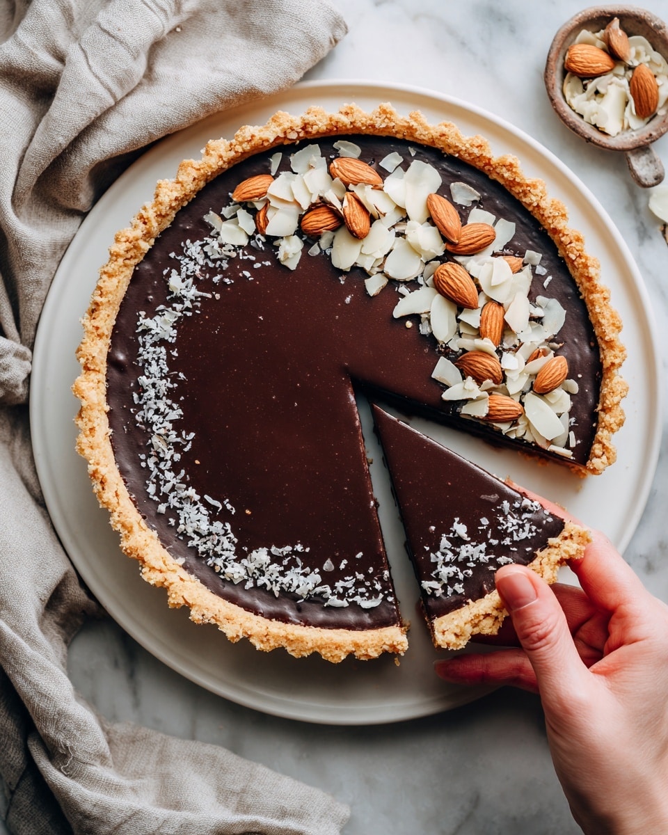 A chocolate tart on a white plate with a slice removed, showing three layers: a crumbly brown crust, a thick, glossy dark chocolate filling, and a topping of sliced almonds and whole almonds arranged in the center over a light dusting of powdered sugar. The tart sits on a white marbled surface, and a woman’s hand is reaching towards the plate. Photo taken with an iphone --ar 4:5 --v 7