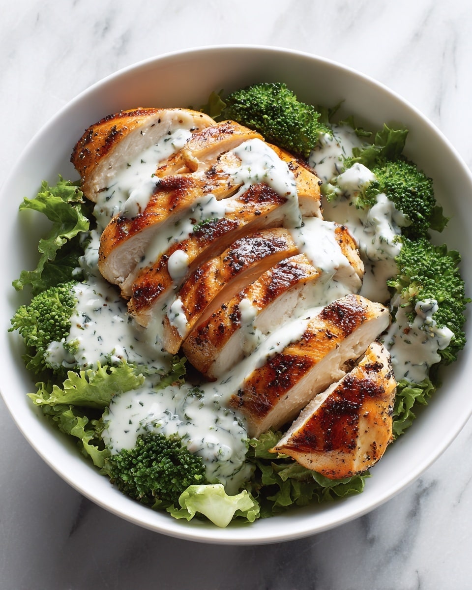 A white bowl holds a dish with two main layers: at the bottom is a bed of green lettuce and bright green broccoli florets arranged around the edges, and on top is a sliced grilled chicken breast with a golden-brown, slightly charred surface. The chicken is sliced into thick pieces fanned out across the greens. A creamy white sauce with specks of green herbs is drizzled generously over the chicken. The background is a white marbled texture. Photo taken with an iphone --ar 4:5 --v 7