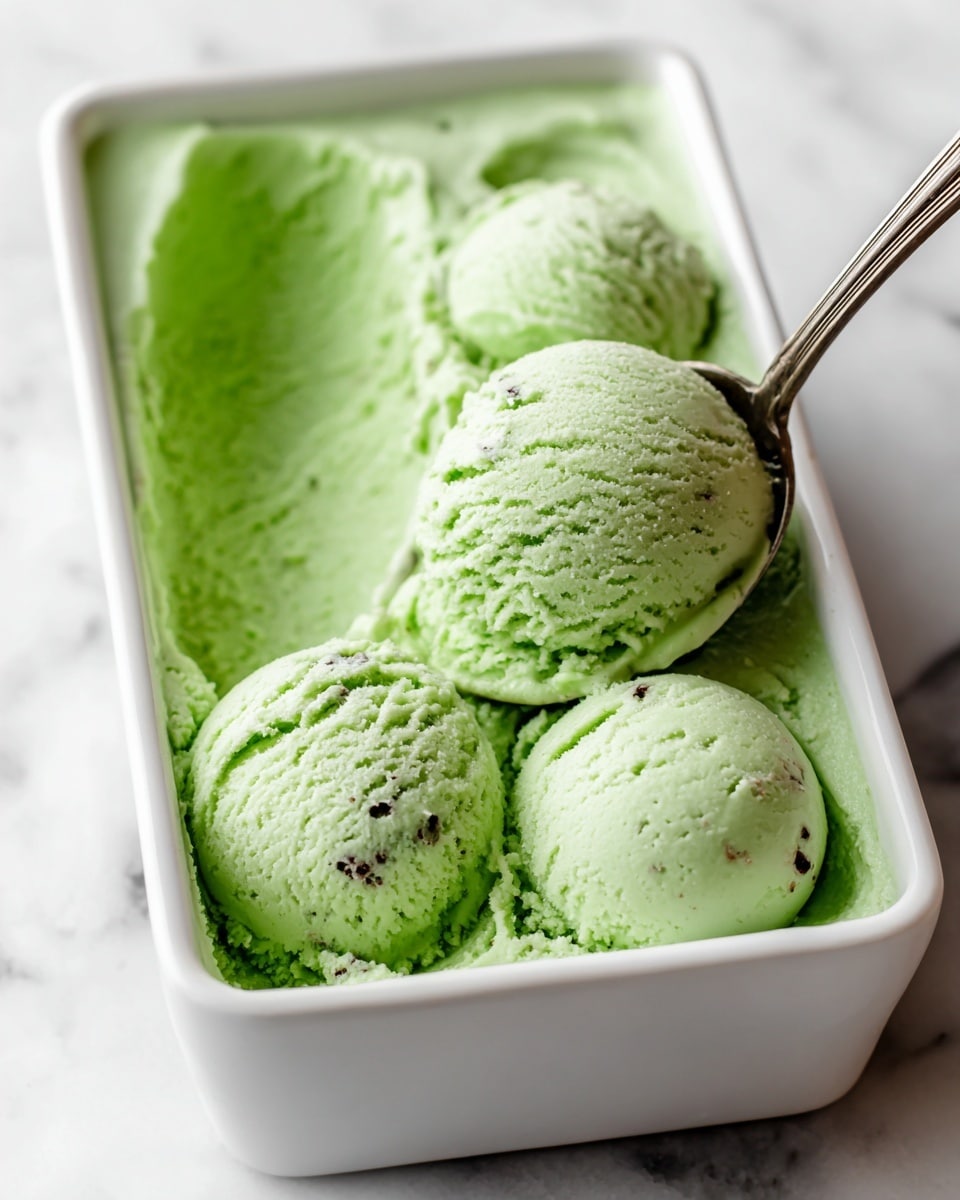 A close-up view of bright green ice cream with a creamy texture and specks of darker green and brown inside, scooped into three round balls sitting in a long white rectangular container filled with the same green ice cream base; a woman’s hand holding a silver spoon is lifting one of the scoops, with the background showing a soft white marbled texture. Photo taken with an iphone --ar 4:5 --v 7