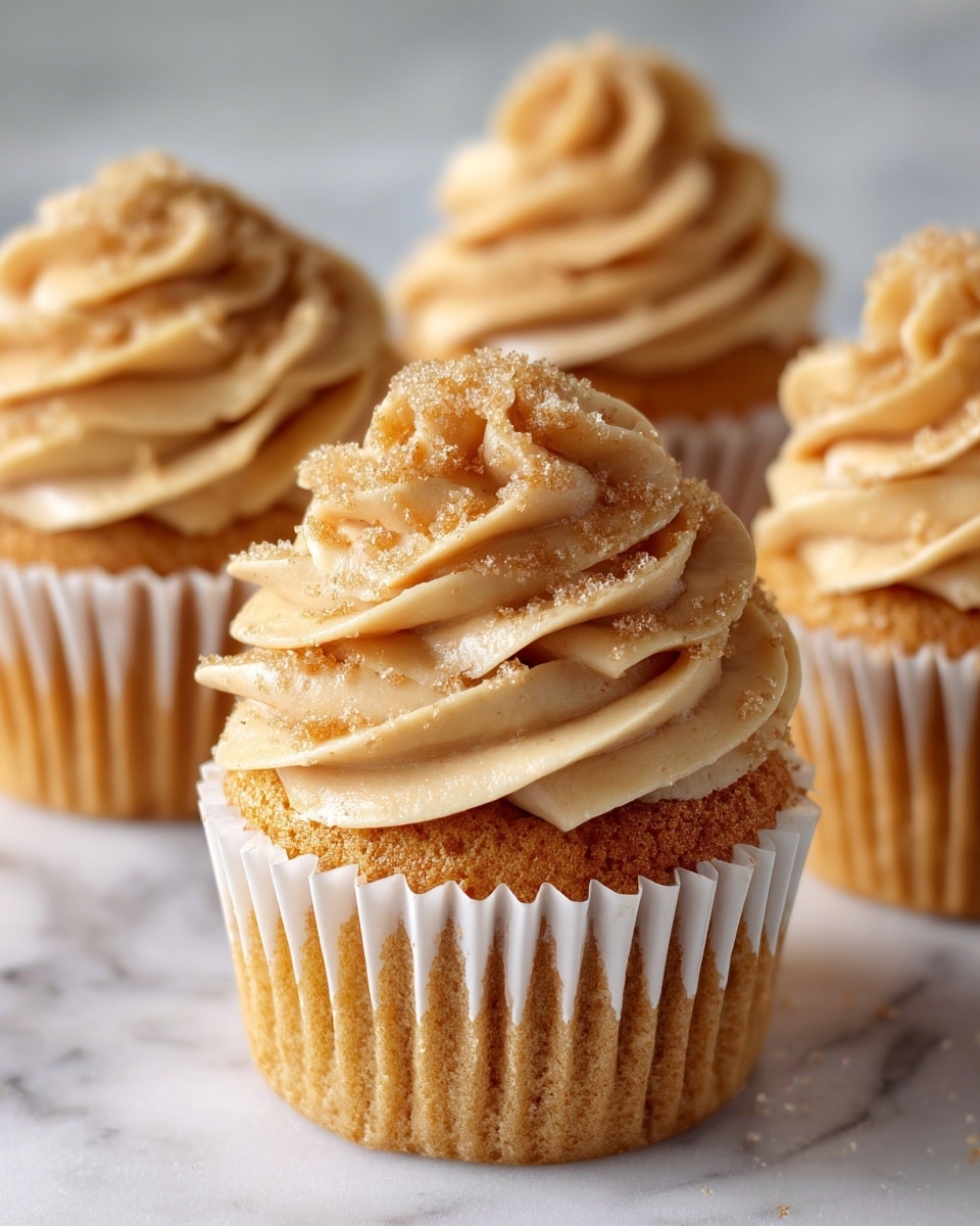 The image shows a close-up of four cupcakes on a white marbled surface. Each cupcake has a single golden brown cake layer at the bottom with a soft, smooth texture. On top, there is one thick swirl of light cream-colored frosting, piped high with a delicate, fluffy texture. The frosting is sprinkled lightly with fine golden crumbs or spices, adding a slight rough texture and a warm color contrast. The cupcakes are in plain light beige paper liners. The focus is on the front cupcake, with the other three blurred softly in the background. Photo taken with an iphone --ar 4:5 --v 7