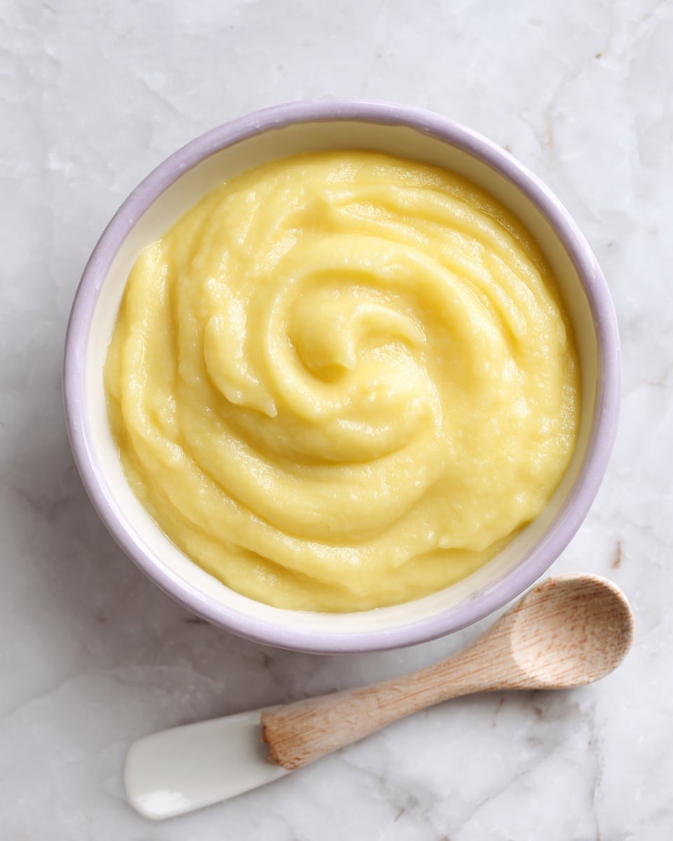 A small white bowl filled with smooth, creamy yellow polenta that has a slightly glossy surface with soft, swirled peaks. The bowl is placed on a white marbled texture with part of a yellow cloth visible on the left side, adding a warm touch to the scene. Photo taken with an iphone --ar 4:5 --v 7