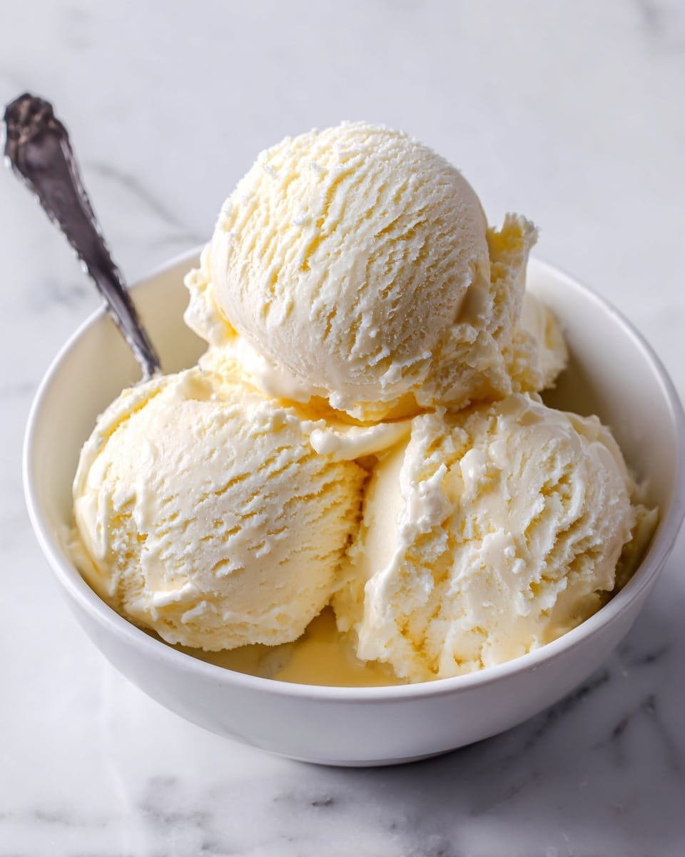 A close-up image shows three scoops of pale yellow ice cream stacked in a small white bowl. The ice cream has a smooth and creamy texture with soft ridges from scooping. A metal spoon is partially visible, resting inside the bowl on the right side. The bowl sits on a white marbled surface that adds a clean and bright background to the image. The lighting highlights the creamy texture and soft color of the ice cream. Photo taken with an iphone --ar 4:5 --v 7