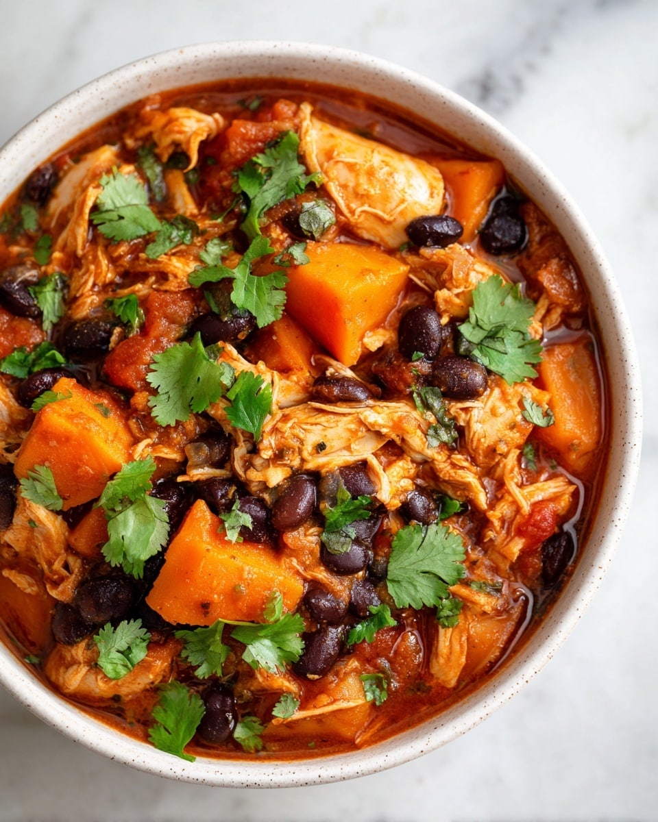 A close-up view of a bowl filled with a thick stew containing large orange cube-shaped sweet potato pieces, shredded light brown chicken, and black beans mixed throughout in a rich, reddish-brown sauce, with small bits of green vegetables and tomatoes visible. The stew is topped with fresh green cilantro leaves scattered over the surface. The bowl is white and has a speckled texture. The scene is set against a white marbled background. photo taken with an iphone --ar 4:5 --v 7