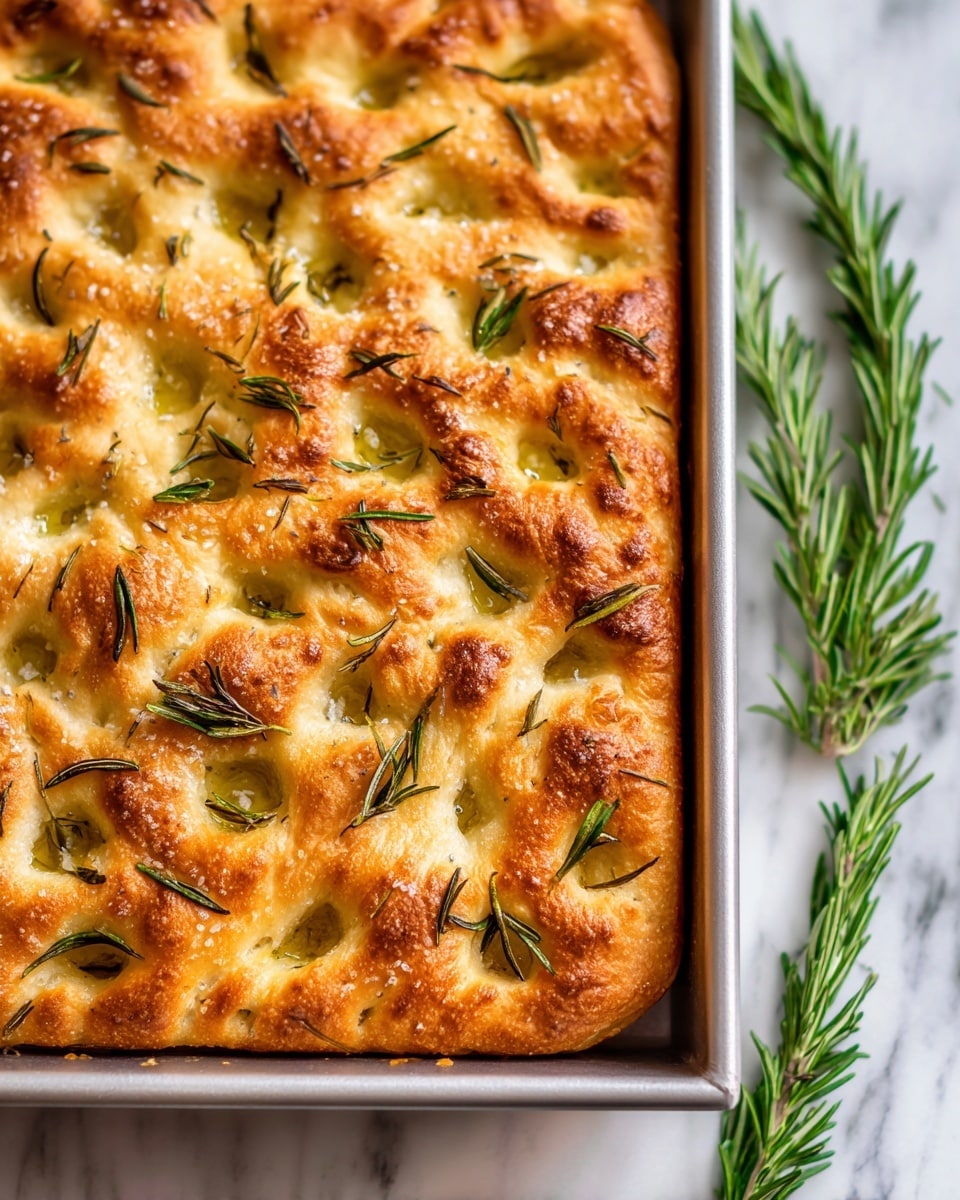 A close-up view of a golden brown focaccia bread in a square metal baking pan. The bread has a slightly puffy texture with many dimples on the surface filled with olive oil. Small green rosemary leaves are scattered evenly across the top, along with coarse sea salt. The bread looks soft inside but has a crisp crust outside. The pan rests on a white marbled surface with some sprigs of fresh rosemary around. Photo taken with an iphone --ar 4:5 --v 7