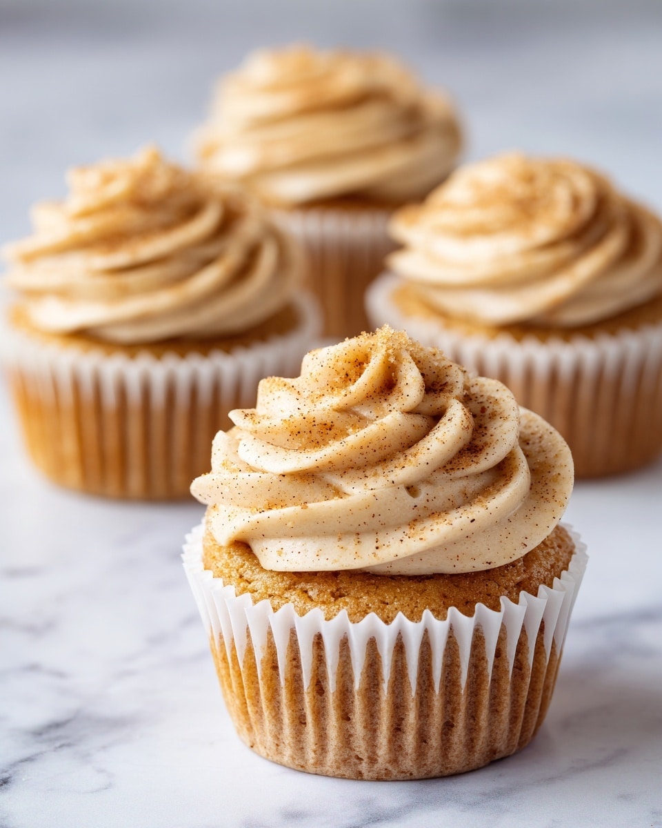 Four golden brown cupcakes with light tan frosting swirled on top, sprinkled with small crumbs or sugar pieces. The cupcakes are in white paper liners, sitting on a white marbled surface. The frosting has a smooth, creamy texture, and each cupcake is round with a slightly domed top. The focus is on the front cupcake, with the others softly blurred in the background. photo taken with an iphone --ar 4:5 --v 7