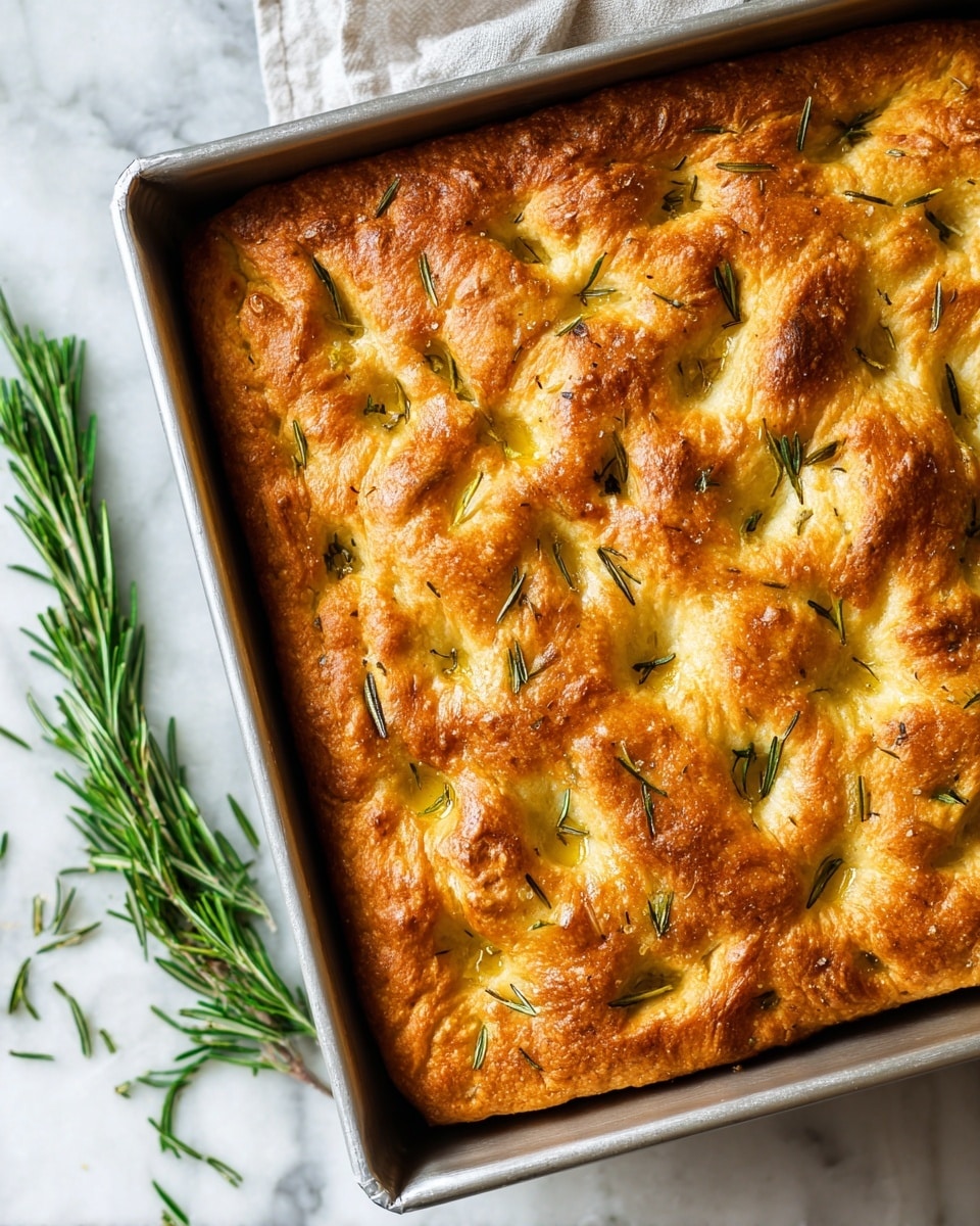 A close-up image of a freshly baked focaccia bread in a square silver baking pan, showing one thick golden brown crust layer with a slightly uneven surface. The bread has many dimples on top, filled with olive oil, sprinkled with coarse sea salt crystals and small green rosemary leaves scattered across. The baking pan rests on a white marbled textured surface, with a few green rosemary sprigs lying nearby. Photo taken with an iphone --ar 4:5 --v 7