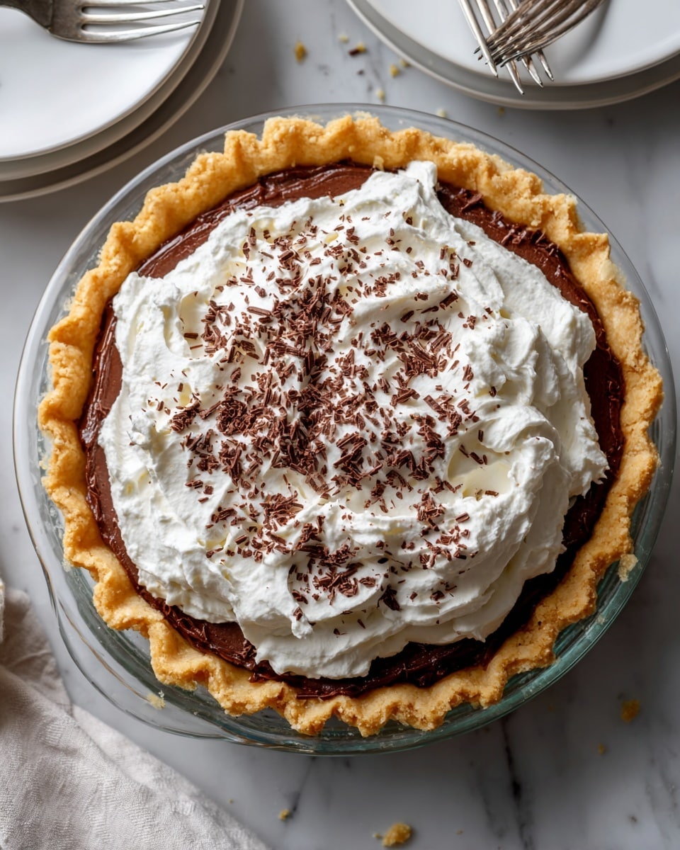A clear glass pie dish holds a three-layer dessert placed on a white marbled surface. The bottom layer is a light brown crust that looks crumbly and thick, lining the edges and base evenly. Above it is a smooth, darker brown chocolate filling that fills most of the pie, topped by a thick, fluffy white whipped cream layer spread evenly with soft curls. Small dark chocolate shavings are sprinkled on top of the whipped cream. To the side, there is a white plate with some crumbs and two forks placed on it, and another empty white plate nearby. Photo taken with an iphone --ar 4:5 --v 7