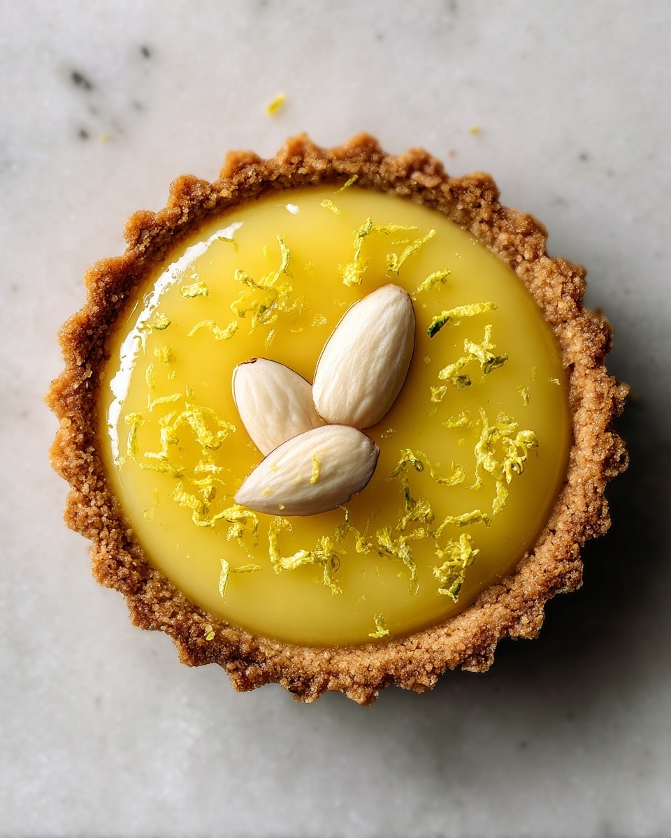 A small tart sits directly on a white marbled texture. The tart has one visible layer of golden, crumbly crust forming a round cup shape. Inside is a smooth, glossy yellow filling with a slightly translucent look. On top of the filling, there are two whole almond nuts placed in a crossed position. Around the tart, small pieces of yellow zest are scattered loosely on the white marbled surface. The lighting is soft, highlighting the textures and colors clearly. photo taken with an iphone --ar 4:5 --v 7